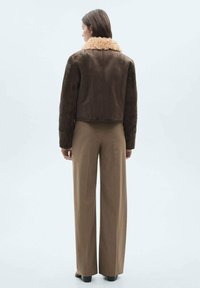 Jachetă scurtă maro cu un guler de blană texturată de culoare bej, purtată cu pantaloni maro cu talie înaltă. Vista din spate evidențiază detalii ale cusăturilor jachetei.