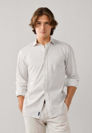 Joven con el pelo castaño de longitud media de pie frente a un fondo liso, vestido con una camisa blanca abotonada y pantalones claros, con las manos en los bolsillos.
