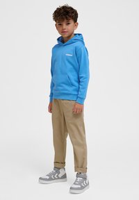 Blauer Kapuzenpullover mit einer Fronttasche und Logo, kombiniert mit beigen Hosen und grauen Turnschuhen. Das Outfit zeichnet sich durch eine lässige Passform und weiches Material aus.