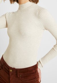 Femme portant un pull à col roulé beige côtelé à manches longues, rentré dans un pantalon en velours côtelé marron taille haute, une main sur la hanche.