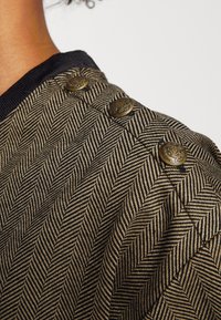 Quiltad jacka i svart och guld med ett fiskbensmönster. Har tre dekorativa knappar på axeln i mässingsfinish.