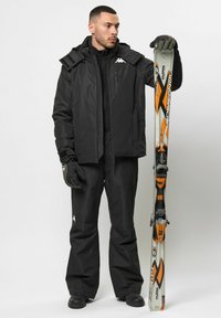 Schwarze wasserdichte Skijacke mit abnehmbarer Kapuze, passende Hose und Handschuhe. Skiausrüstung mit orangefarbenen Akzenten und Bindungen.
