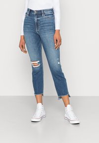 Jeans de denim azul de cintura alta com dois rasgos nos joelhos e bainhas desfiadas, combinados com sapatilhas brancas de cano alto e uma blusa justinha de manga longa.