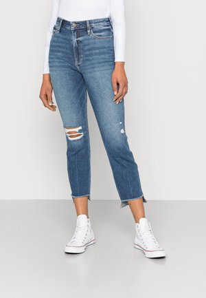 Jeans de denim azul de cintura alta com dois rasgos nos joelhos e bainhas desfiadas, combinados com sapatilhas brancas de cano alto e uma blusa justinha de manga longa.