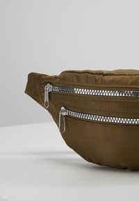 Sac banane en tissu marron avec deux poches zippées en métal argenté sur un fond gris et blanc uni.
