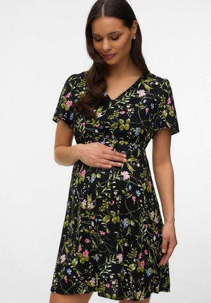 Femme aux longs cheveux foncés portant une robe noire à manches courtes à motif floral, tenant délicatement son ventre de femme enceinte, debout devant un fond uni.