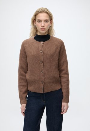 Lyshåret kvinde iført sølv hoop øreringe, brun knappet cardigan over sort top og mørkeblå jeans, stående mod en hvid baggrund.