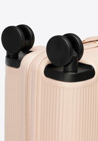 Beige Hartschalentrolley mit vertikalen Rillen, ausgestattet mit zwei Sets von schwarzen Doppel-Spinnrädern in den oberen Ecken.