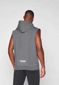 Bezrukávová šedá mikina vyrobená z jemného materiálu, s veľkou kapucňou a bielym logom "VENUM" na dolnej časti chrbta. Jednoduchý dizajn, bez vzorov.