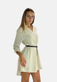 Beige overhemdjurk met lange mouwen, een kraag en een zwarte ceintuur in de taille. De stof heeft een gestructureerde uitstraling en valt soepel naar een gebogen zoom.