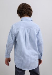 Ljusblå skjorta med knappar och vertikala vita ränder, långärmad och med rundad nederkant. Har en klassisk krage och knäppta manschetter.