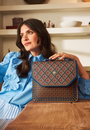 Vrouw in een blauwe blouse die haar arm op een houten tafel laat rusten en een handtas vasthoudt met bloem- en geometrische patronen.