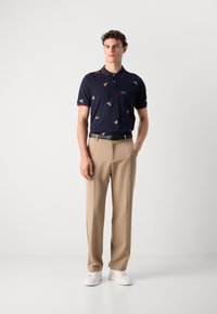 Marineblå poloskjorte med små fuglemønstre, rød kragebesetning, beige bukser og hvite joggesko; klassisk passform med korte ermer og knapper foran.