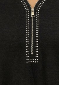 Zwarte pullover met een centrale rits, versierd met zilveren studs die in twee verticale lijnen langs de halslijn zijn gerangschikt. Zachte stoffen textuur.