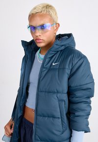 Πάχη navy μπλούζα με κουκούλα και φερμουάρ, με το λευκό λογότυπο της Nike. Φορεμένη με μια ανοιχτού μπλε μπλούζα. Ορατά ασημένια αξεσουάρ.