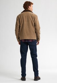 Giacca in corduroy marrone con un colletto scuro e peloso, indossata sopra una camicia a quadri. Pantaloni in denim blu e scarpe marroni visibili da dietro.