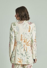 Blazer bordado en lino crema con patrones florales multicolores. Presenta un diseño ajustado, botones en los puños y una textura suave.
