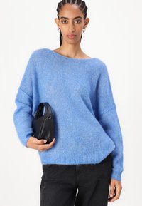 Γαλάζιο φουσκωτό πουλόβερ με φαρδύ λαιμό και φαρδιά εφαρμογή, συνδυασμένο με μια μικρή μαύρη τσάντα. Η υφή του φαίνεται μαλακή και πλεκτή με μακριά μανίκια.