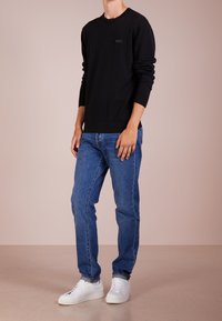 Hombre vistiendo una camisa de manga larga negra, jeans azules y zapatillas blancas, de pie sobre un suelo beige contra una pared de color beige claro.