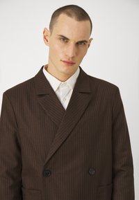 Dobbeltbrystet brun blazer med pynte-striber lavet af tekstureret stof, med smalle klipper, to sorte knapper og en forlommer.