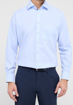Uomo che indossa una camicia azzurro chiaro a maniche lunghe con bottoni sul davanti e tasca, abbinata a pantaloni blu scuro e cintura nera.