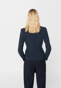 Blazer bleu marine à épaules structurées, manches longues et coupe ajustée. Texture lisse avec un style minimaliste. Associé à un pantalon rayé.