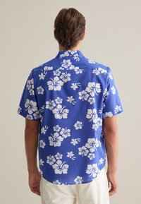 Camisa de manga corta azul con patrón floral de hibisco blanco. Tejido ligero, cuello y dobladillo curvado en la parte posterior.