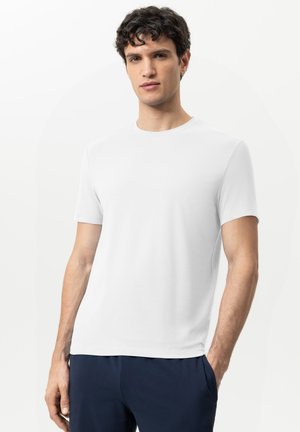 Junger Mann mit kurzen lockigen Haaren, der ein schlichtes weißes kurzärmeliges T-Shirt und dunkelblaue Hose trägt, steht mit einer Hand in der Tasche.