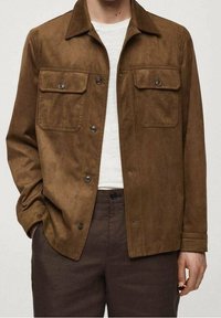 Veste en daim marron avec deux poches poitrine, fermeture à boutons et col classique. Portée sur une chemise blanche, associée à un pantalon foncé.