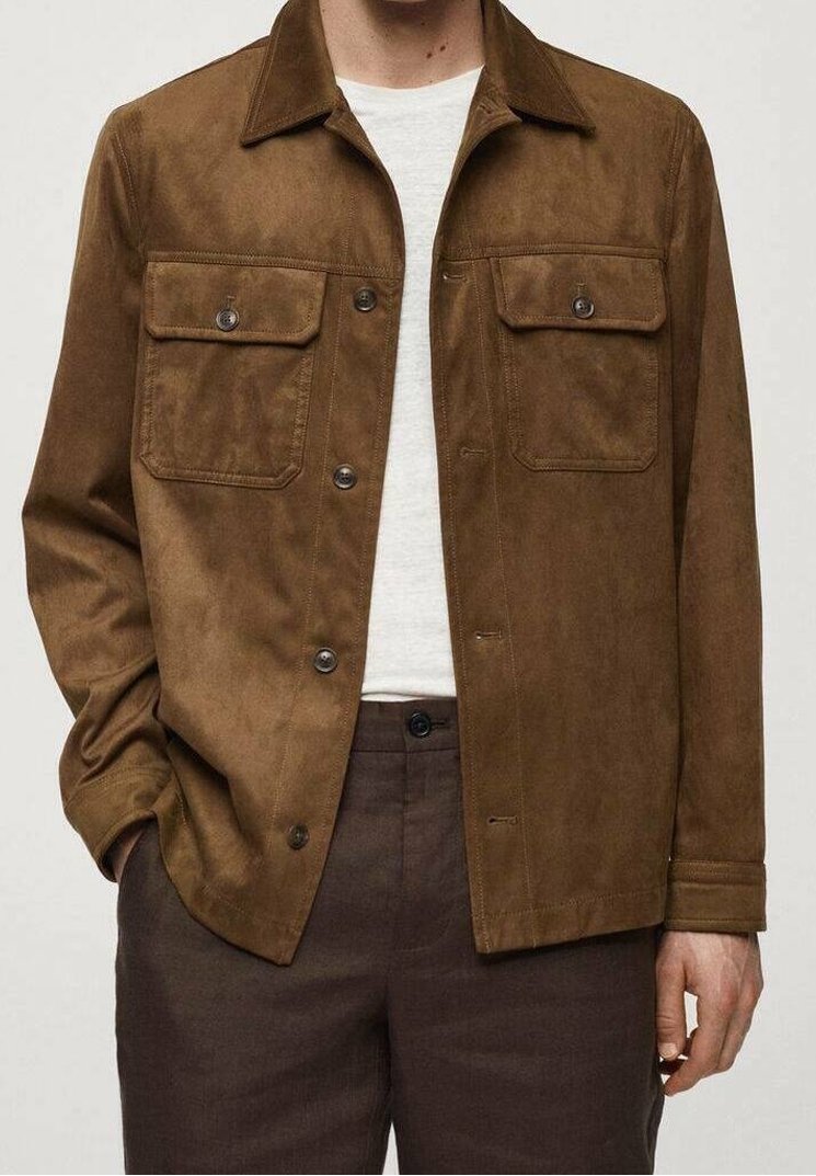 Veste en daim marron avec deux poches poitrine, fermeture à boutons et col classique. Portée sur une chemise blanche, associée à un pantalon foncé.