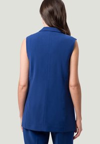 Gilet blu senza maniche con colletto sartoriale, caratterizzato da una texture liscia e orlo dritto, visto da dietro.