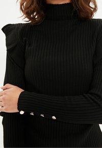 Czarny sweter z prążkowanej dzianiny z golfem, z falbanami na ramionach i dekoracyjnymi złotymi guzikami na mankietach. Miękka faktura, dopasowany krój.