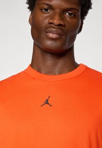 Narancssárga pamut póló kerek nyakkal, fekete Jumpman logóval a mellkason. Sima textúra, laza stílus.
