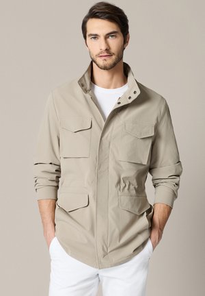 Mężczyzna ubrany w beżową kurtkę roboczą z czterema kieszeniami z patkami i białe spodnie, stoi z rękami w kieszeniach na neutralnym tle.