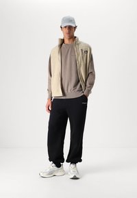 Beige fleece vest over a grey sweatshirt, paired with black sweatpants and white shoes. Grey cap completes the casual outfit.