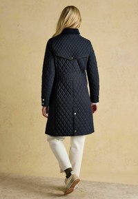 Námornícky prešívaný kabát s golierom, detalom na chrbte a cvočkami. V kombinácii s krémovými nohavicami a svetlými teniskami.