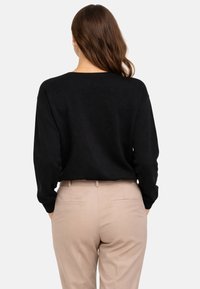 Femme aux longs cheveux bruns portant un pull noir et un pantalon beige, debout les mains dans les poches, vue de dos.