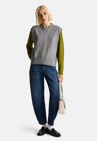 Šedý bezrukávový svetr přes zelenou dlouhouzandálovou košili, doplněný modrými volnými džíny a černými mokasínami. Drží malou bílou kabelku.