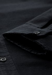 Colletto di camicia nera a trama con chiusura a bottone e cuciture visibili. Il tessuto presenta un sottile motivo intrecciato. Finitura morbida e liscia.