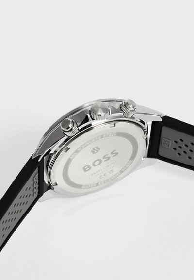 Dos de montre-bracelet en acier inoxydable argenté avec bracelet en caoutchouc noir perforé et trois boutons de réglage latéraux.