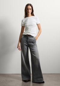 Witte geribde short-sleeve shirt gecombineerd met wijde grijze jeans met een gladde textuur en een voorkeuzevak. Een bruine riem maakt de look compleet.