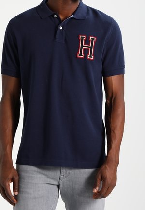 Homme portant un polo bleu marine avec un "H" rouge et blanc brodé sur la poitrine gauche et un jean gris clair.