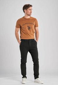 Ruskea T-paita, jossa on U.S. Polo Assn. -grafiikka, mustat housut ja valkoiset lenkkarit. Housuissa on sivutaskut ja napitettu etu.