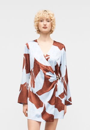 Blond persoon die een lange mouwen mini-jurk met lichtblauw en roestkleurig abstract bloemmotief draagt, staand tegen een effen achtergrond.