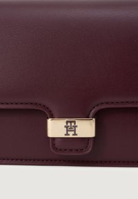 Πορτοφόλι σε μπορντό δερμάτινο χρώμα με υφή, που διαθέτει κούμπωμα σε χρυσό τόνο και ανάγλυφο λογότυπο. Συμπαγής ορθογώνιος σχήμα.