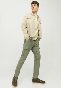 Chaqueta de mezclilla beige, pantalones cargo verde claro con bolsillos laterales y botas marrones con cordones. El conjunto incluye una camiseta de tie-dye ligera debajo.