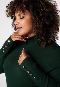 Donkergroene coltrui met geribbelde textuur en gouden knoopaccenten op de mouwen. Hand van het model raakt licht de halslijn.