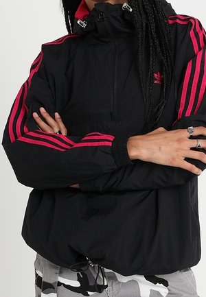 Personne aux longs cheveux tressés portant une veste Adidas noire avec des bandes rouges, bras croisés, et un pantalon camouflage.