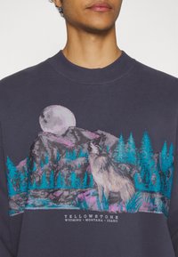 Granatowa bluza z grafiką wilka i gór w żywych odcieniach turkusu i różu, z napisem "YELLOWSTONE, WYOMING, MONTANA, IDAHO".