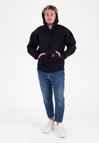 Giovane con le mani in tasca, che indossa una felpa nera con cerniera e cappuccio alzato, jeans blu e sneakers bianche, in piedi su uno sfondo bianco.
